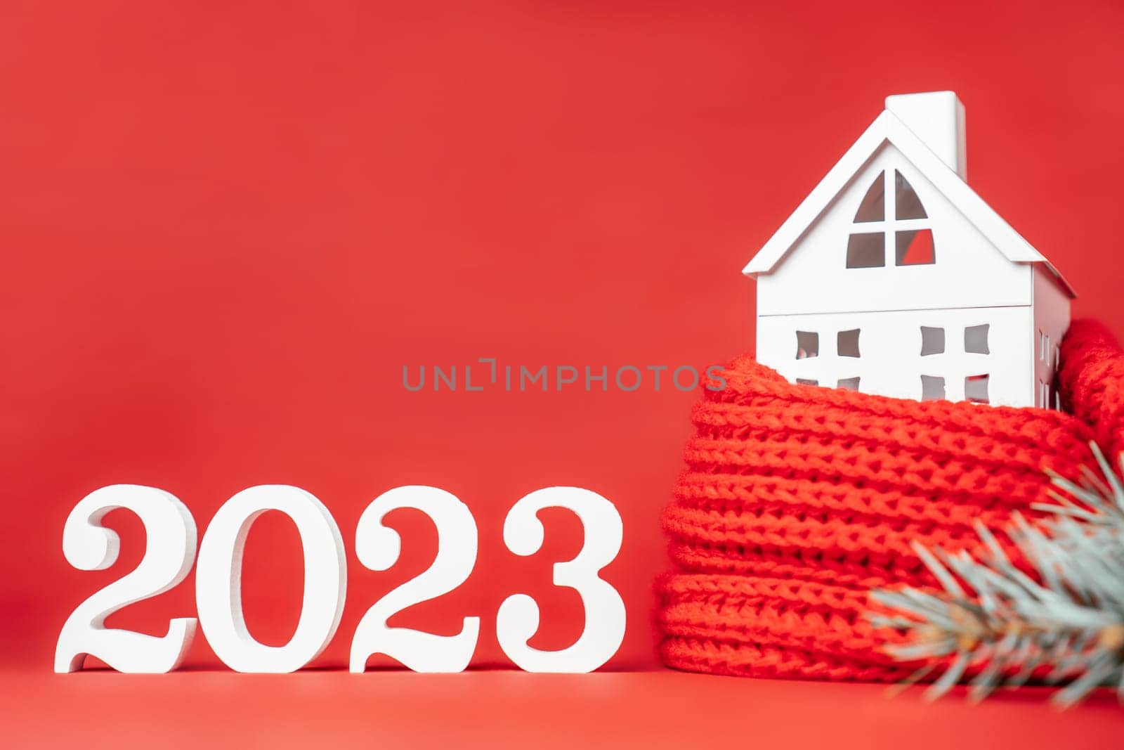 Happy New Year 2023. White wooden numbers 2023 stand with white toy house wrapped in red scarf and Christmas tree branch on red background. Merry Christmas. Copy space