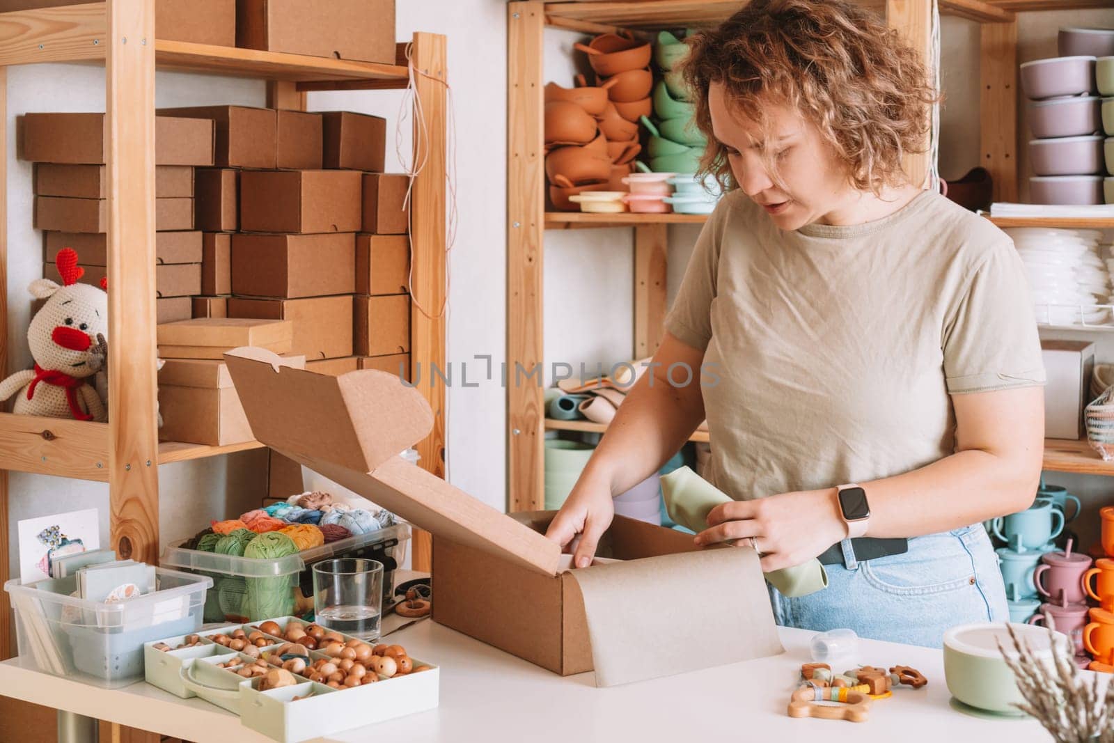 Smiling concentrated curly woman packing and wrapping shipment with silicone baby dish in cardboard box for delivery. Wooden rattle toy, trinket decoration nearby on desk. Workshop store, work at home
