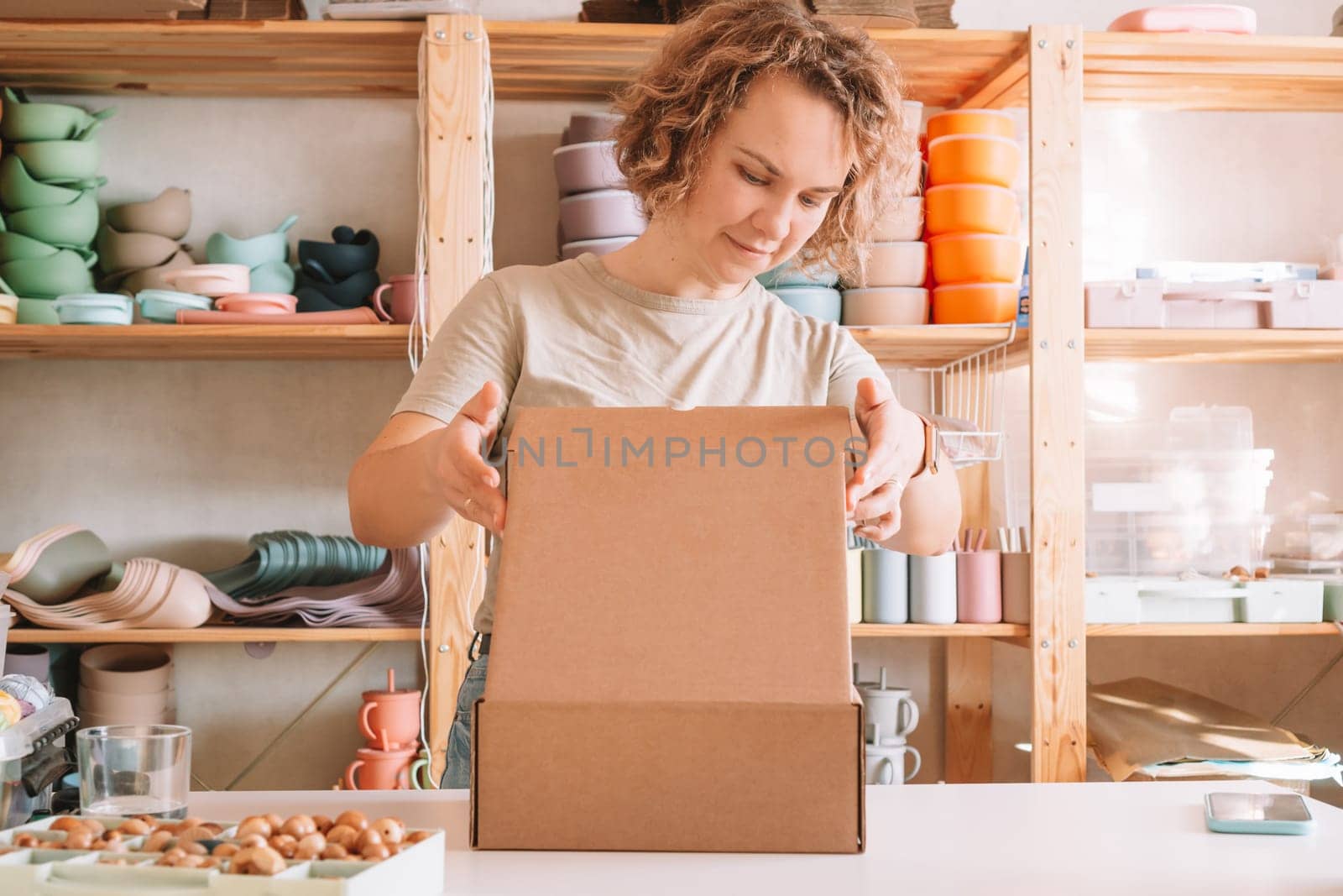 Smiling blond curly woman packing and wrapping shipment, silicone baby dishes in cardboard delivery box. Brown wooden beads in container nearby. Workshop store, small business