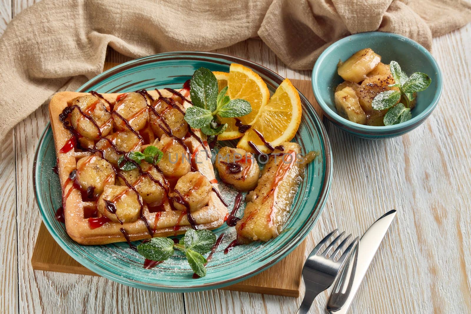 Delicious waffles with caramelized banana, sliced orange, mint, jam and sweet sauce on plate on light wooden background. Top view. Table setting. Dessert. Serving food