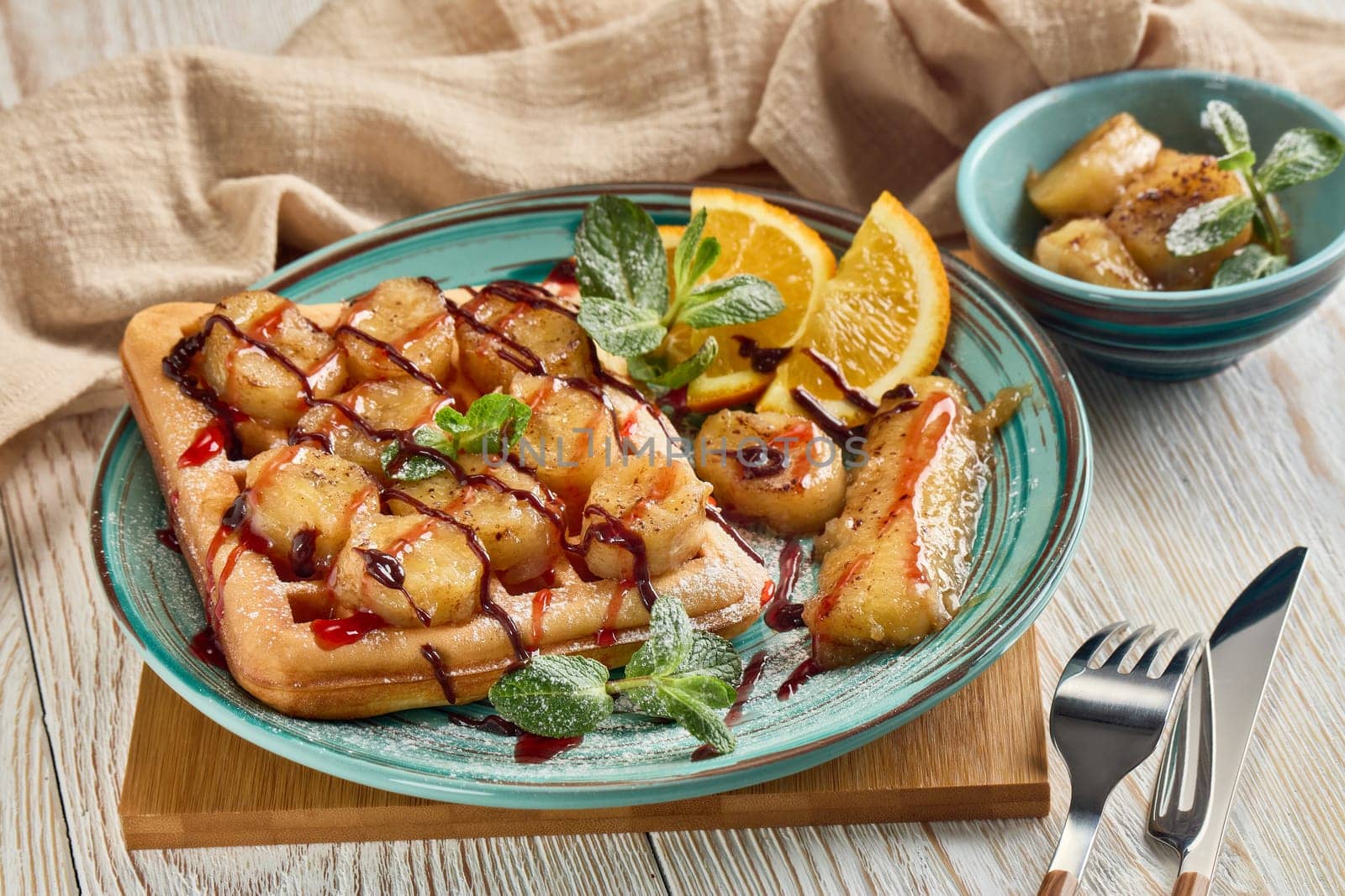 Delicious waffles with caramelized banana, sliced orange, mint, jam and sweet sauce on plate on light wooden background. Table setting. Dessert. Serving food