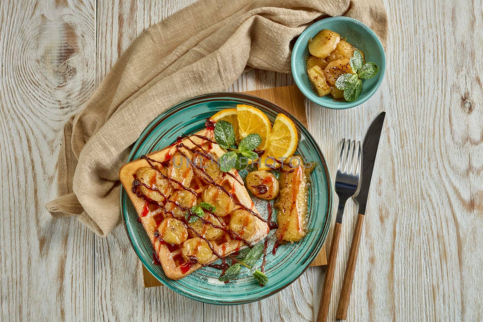 Delicious waffles with caramelized banana, sliced orange, mint, jam and sweet sauce on plate on light wooden background. Top view. Table setting. Dessert. Serving food