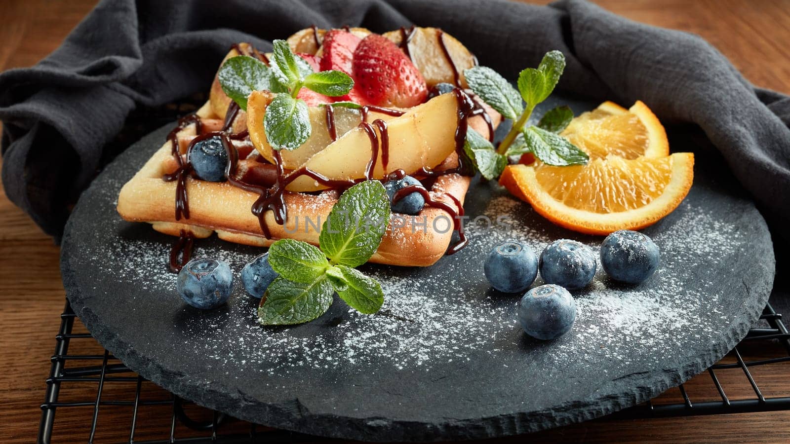 Delicious waffles with caramelized pear, berries, mint, powdered sugar and sweet sauce on black serving board on dark brown background. Dessert. Serving food. Table setting