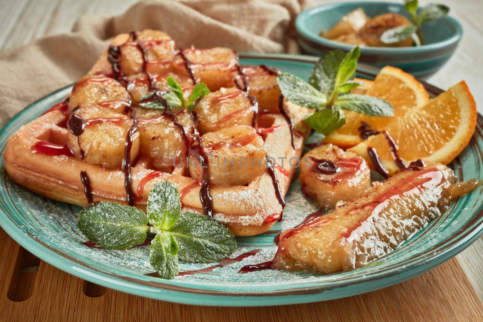 Delicious waffles with caramelized banana, sliced orange, mint, jam and sweet sauce on plate on light wooden background. Table setting. Dessert. Serving food