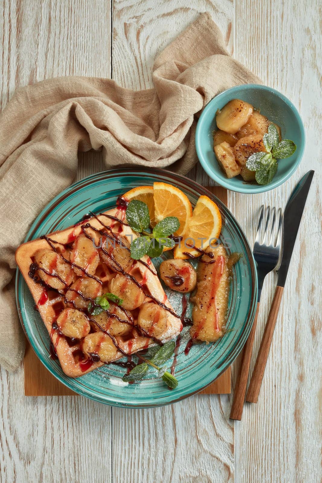 Delicious waffles with caramelized banana, sliced orange, mint, jam and sweet sauce on plate on light wooden background. Top view. Table setting. Dessert. Serving food