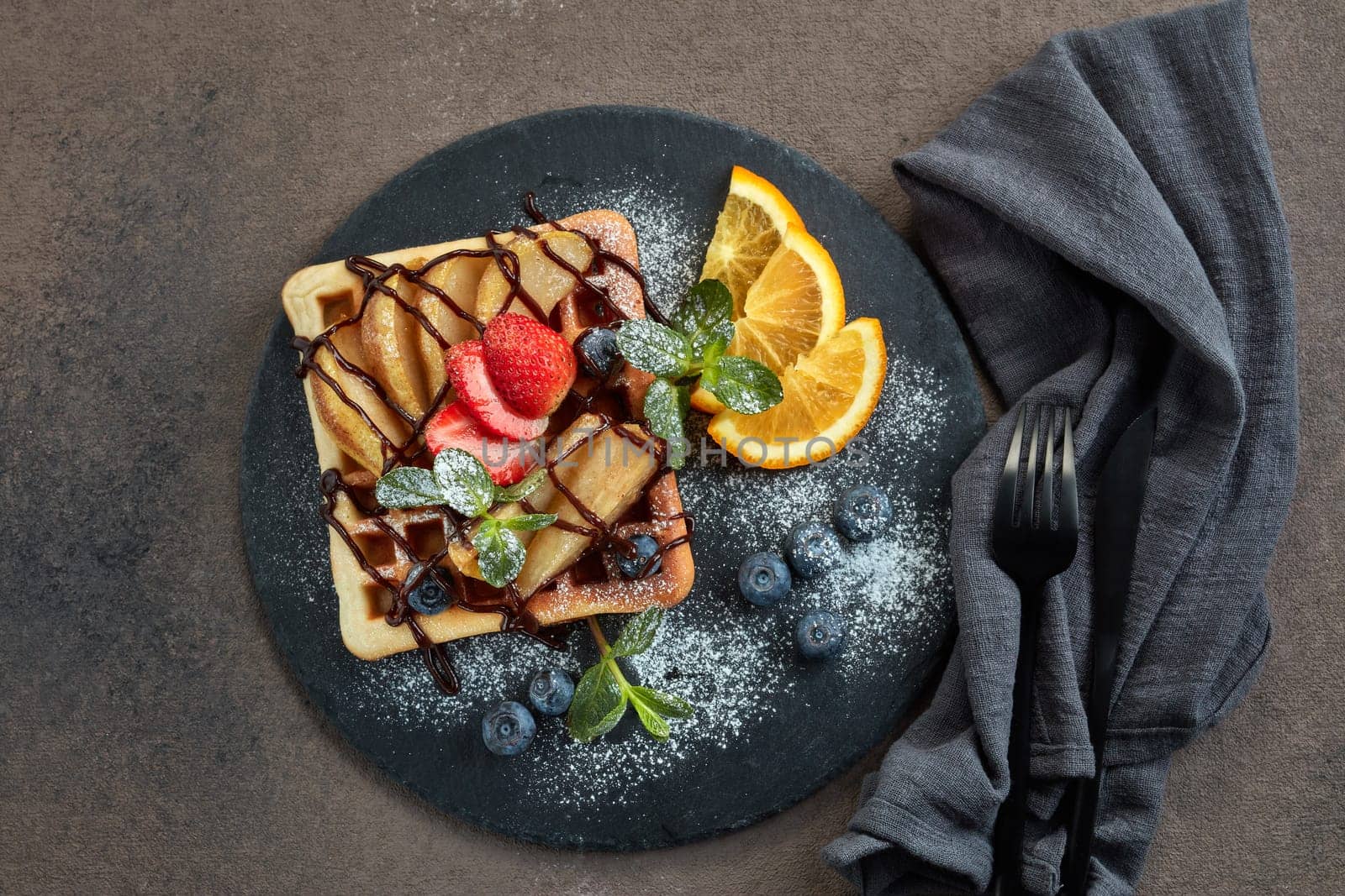 Delicious waffles with caramelized pear, berries, mint, powdered sugar and sweet sauce on black serving board on dark brown background. Dessert. Serving food. Top view