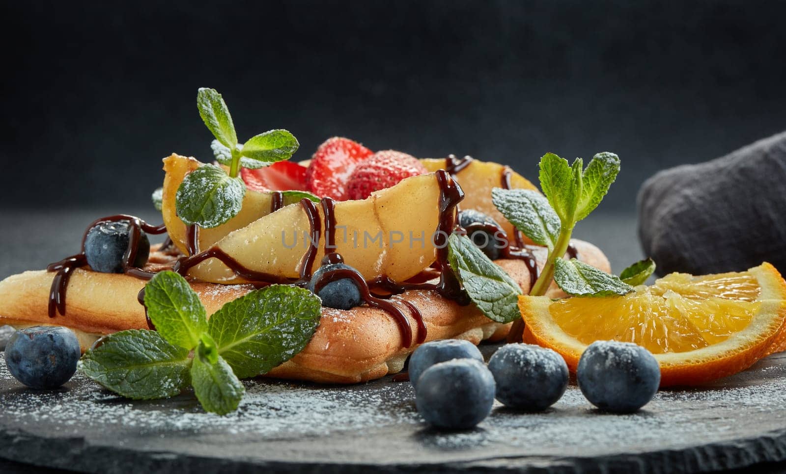 Delicious waffles with caramelized pear, berries, mint, powdered sugar and sweet sauce on black serving board on grey background. Dessert. Serving food. Table setting