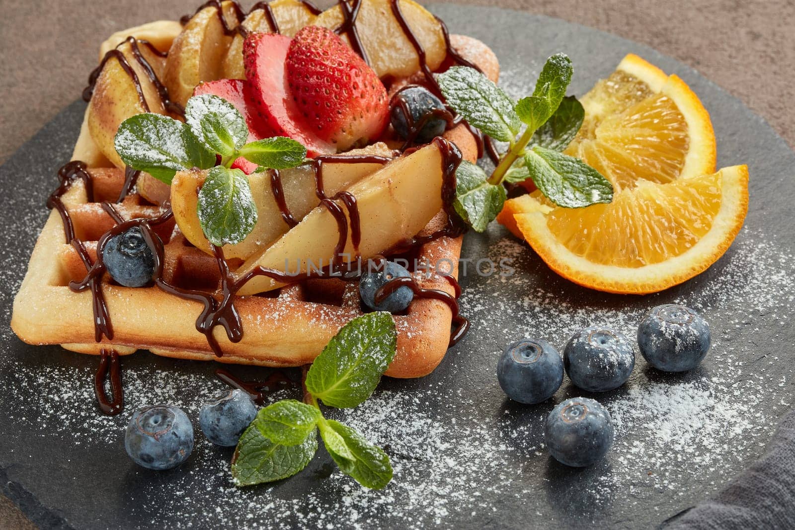Delicious waffles with caramelized pear, berries, mint, powdered sugar and sweet sauce on black serving board on brown background. Dessert. Serving food. Table setting