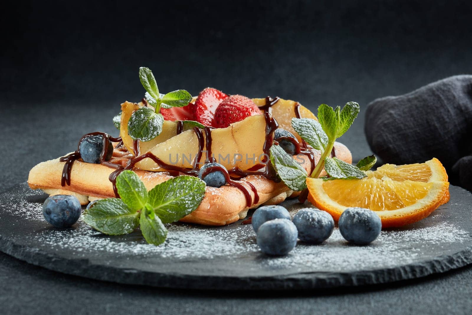 Delicious waffles with caramelized pear, berries, mint, powdered sugar and sweet sauce on black serving board on grey background. Dessert. Serving food. Table setting
