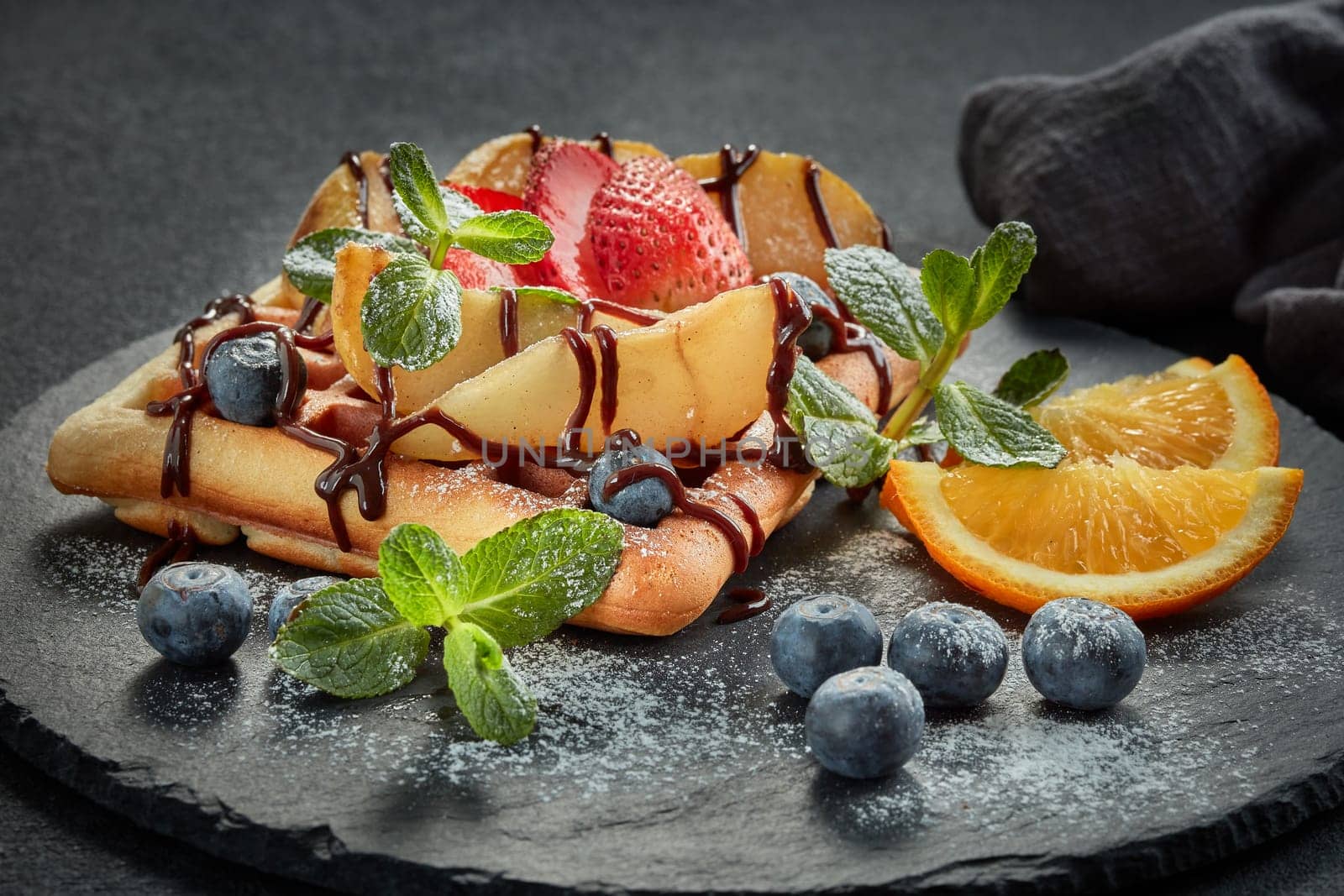 Delicious waffles with caramelized pear, berries, mint, powdered sugar and sweet sauce on black serving board on grey background. Dessert. Serving food. Table setting
