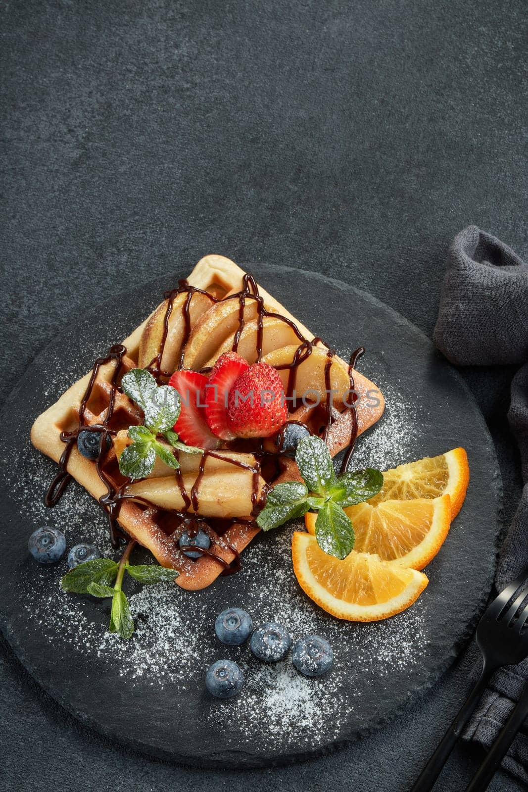 Delicious waffles with caramelized pear, berries, mint, powdered sugar and sweet sauce on black serving board on grey background. Dessert. Serving food. Table setting