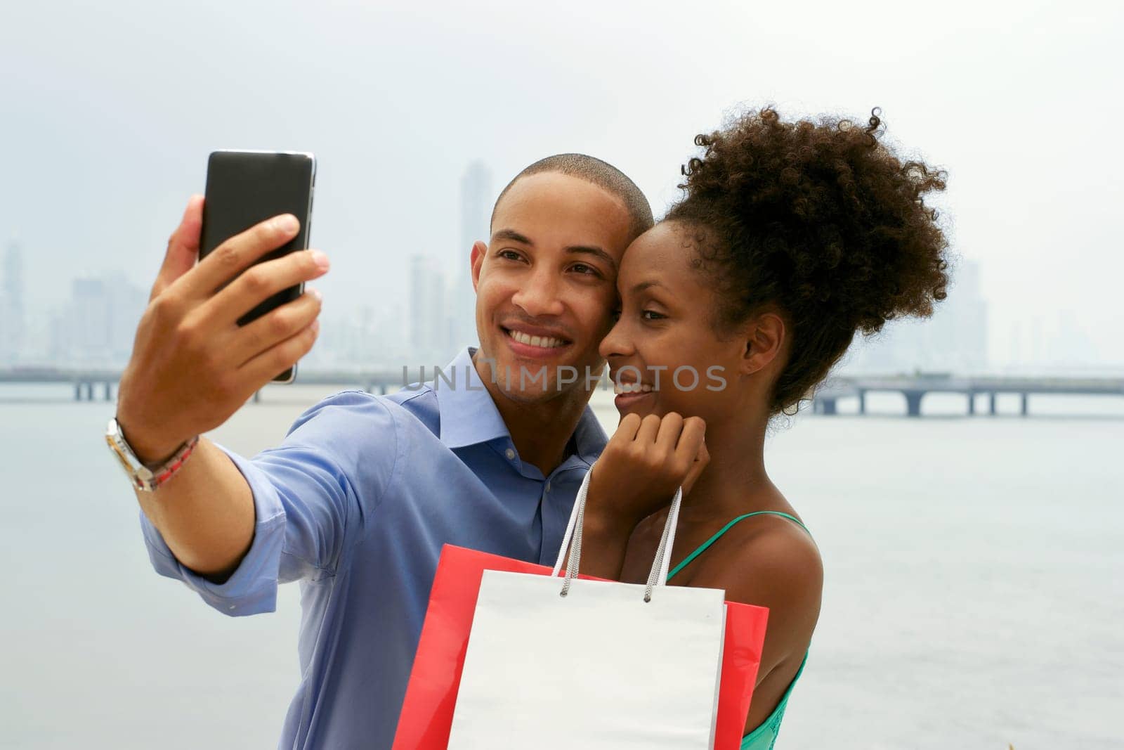 Black tourist heterosexual couple in Casco Antiguo Panama City with shopping bags. The man takes a selfie with his girlfiend and shopping bags with skyline in background