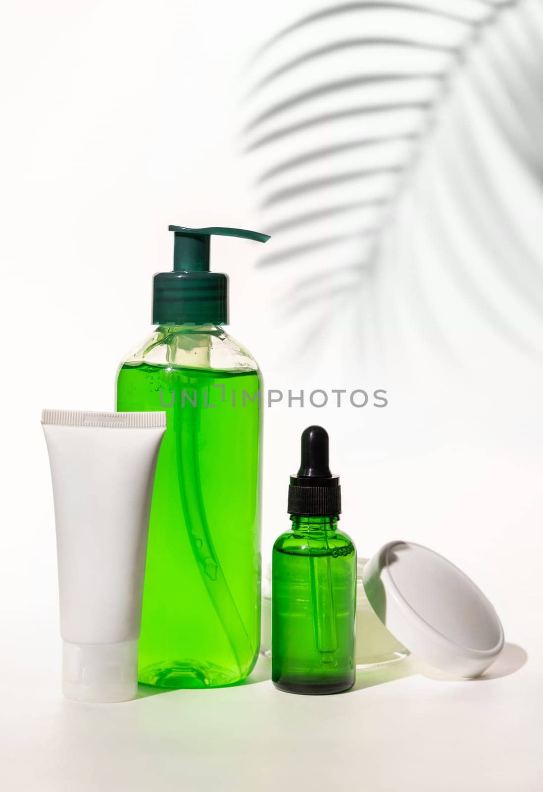 Refillable Cosmetic bottles, jar and tube with green liquid on white, Palm leaf hard shadows, close up, mockup. Skincare beauty products. Natural cosmetics with aloe vera..