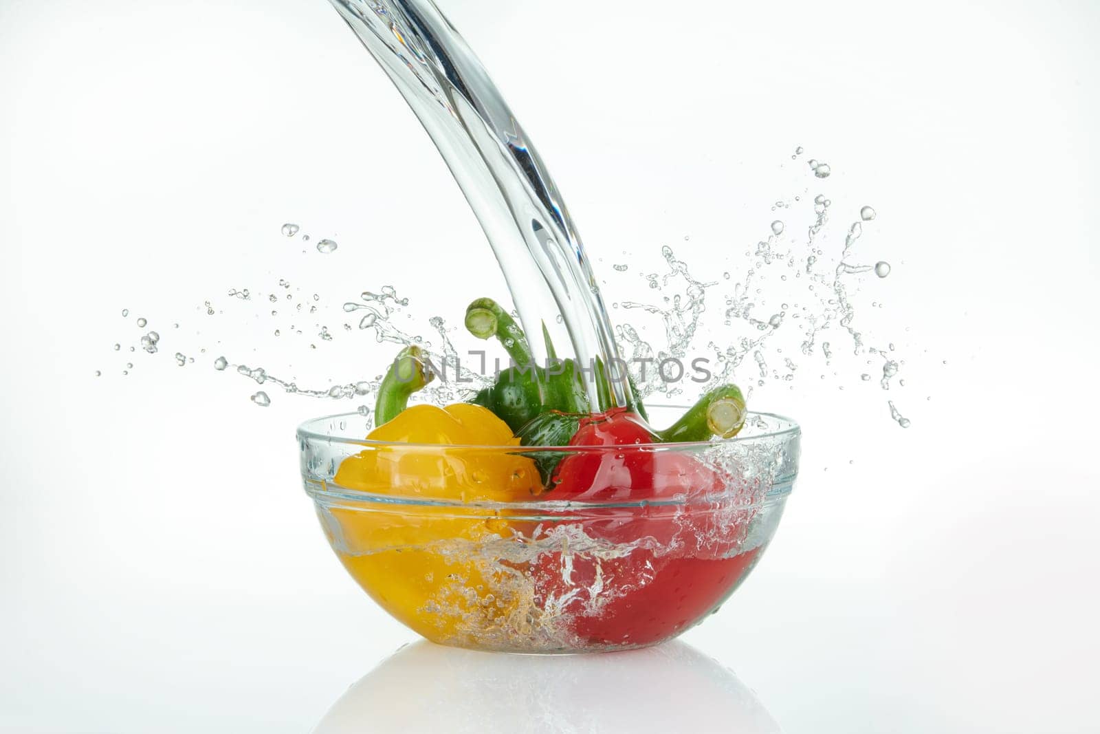 waterfall pouring onto a glass bowl with multicolored peppers with splashes on white background.