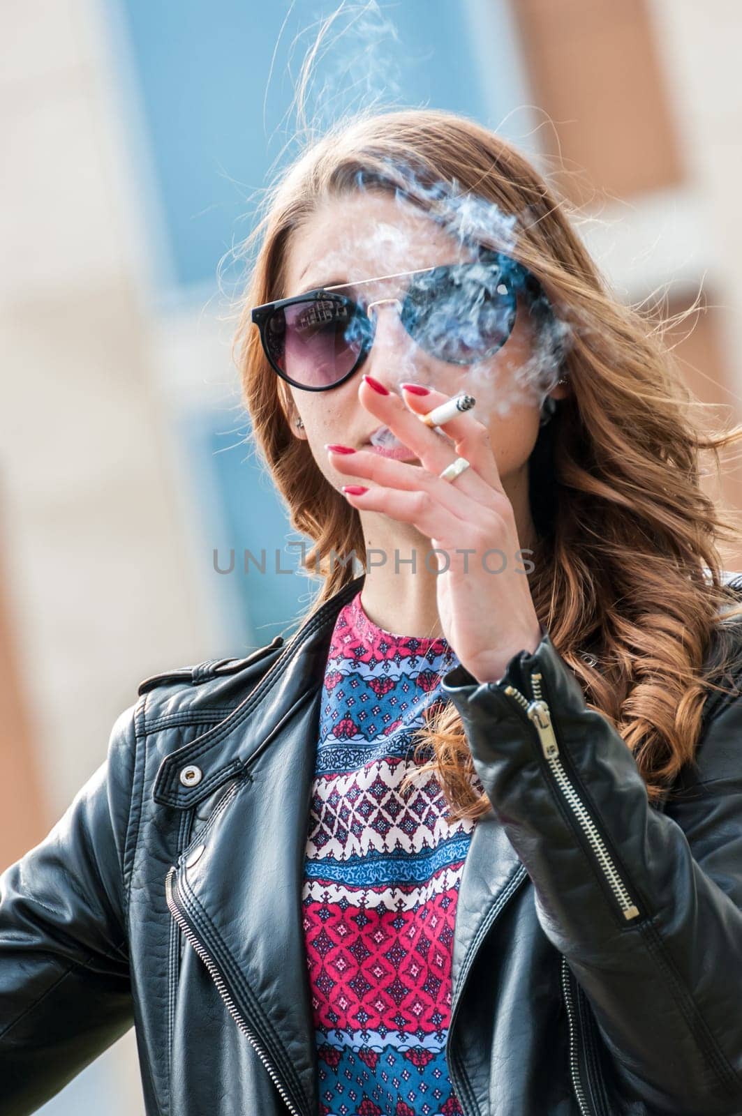 Beautiful curly brunette young Woman smoking a cigarette in front of a building. Enjoying life. The concept of an unhealthy lifestyle.