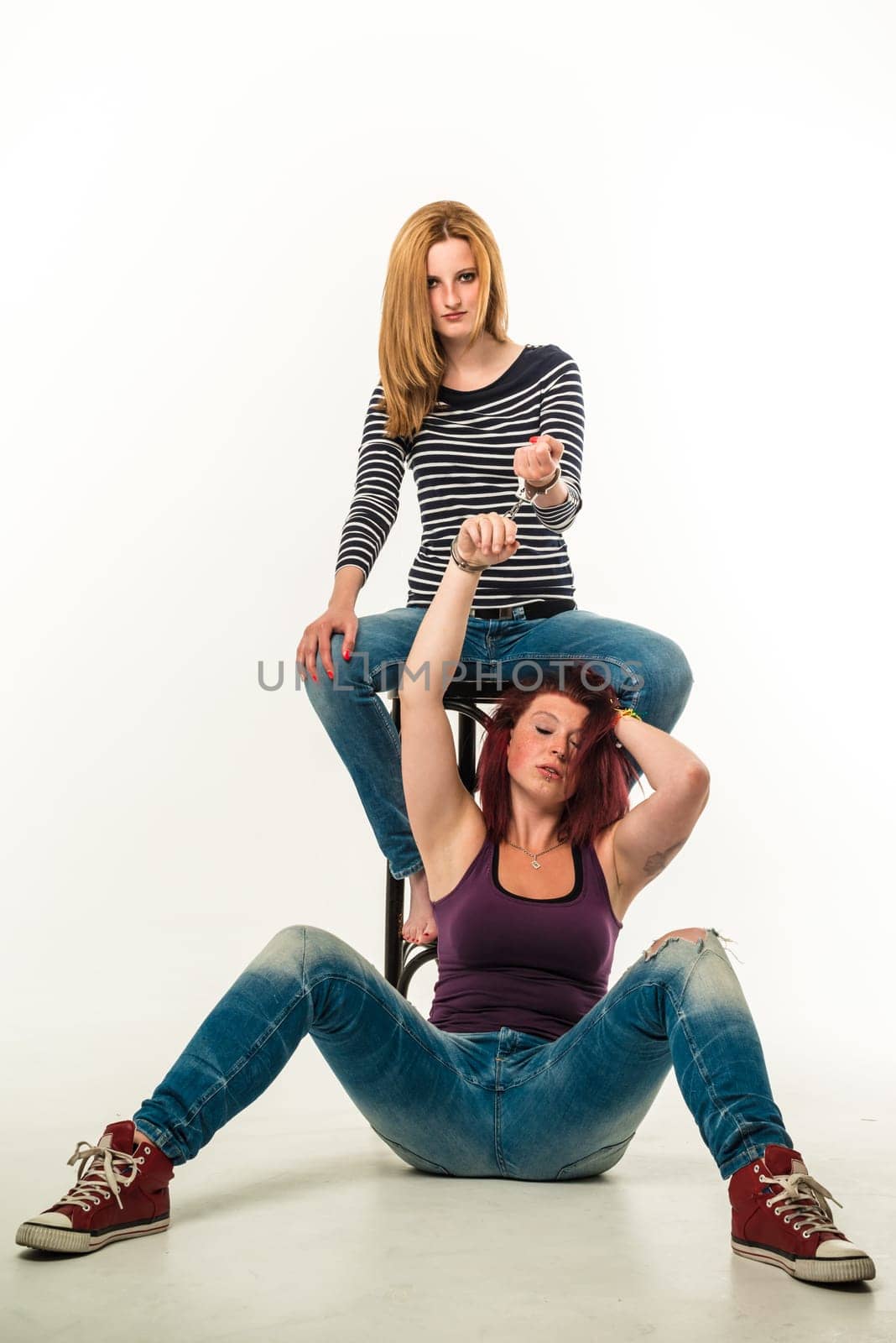 Two angry women connected by a pair of handcuffs wearing jeans, blond and red-haired, barefoot and red chucks, sitting over white background