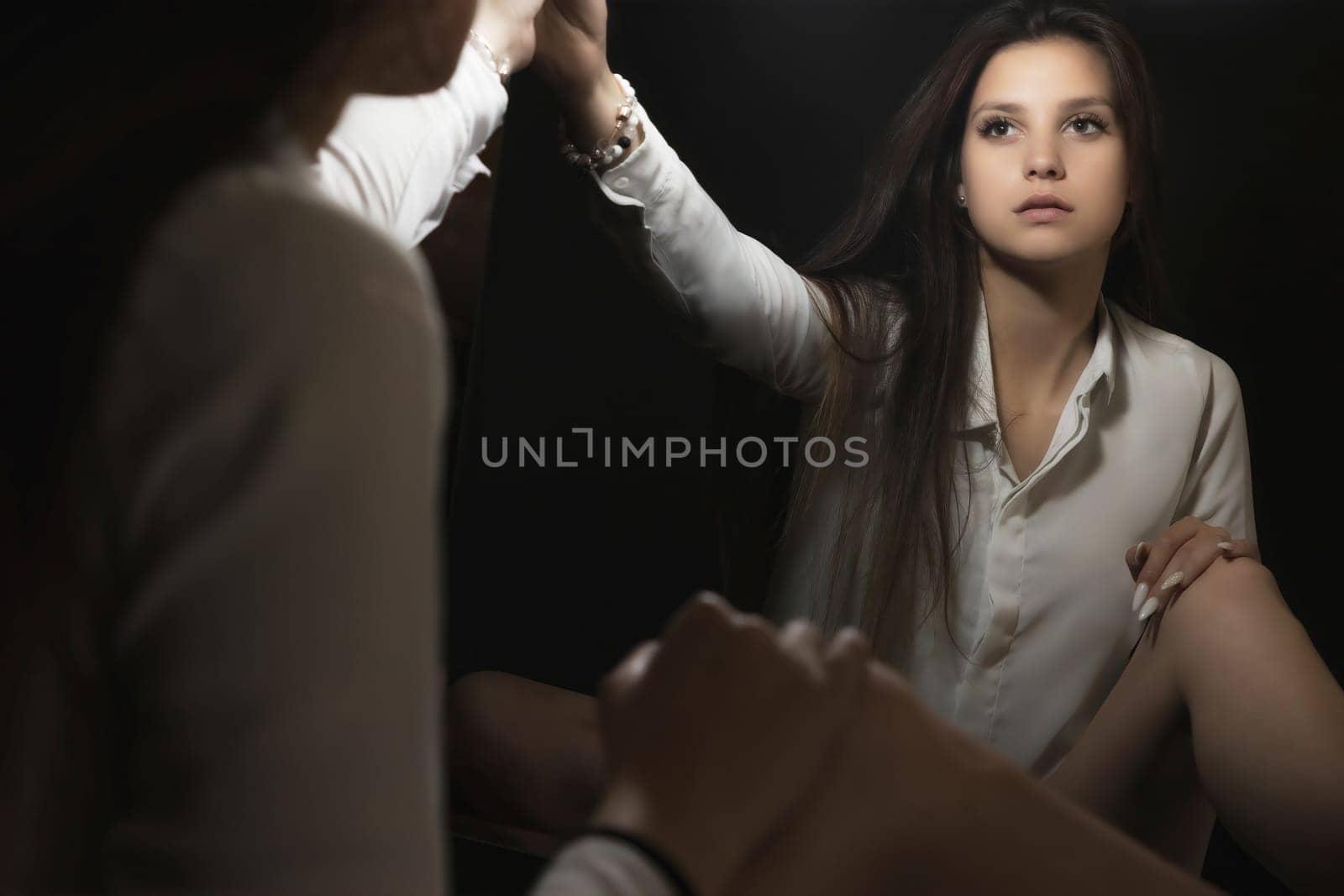 Low key portrait of young woman looking at her reflection in the mirror in a beam of light. Horizontally.