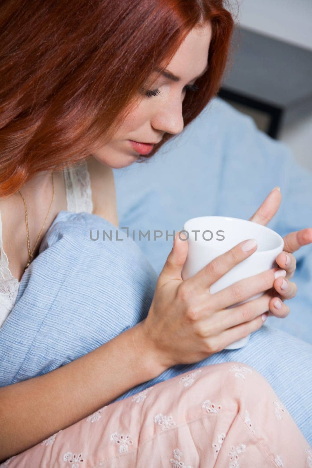 Young red-haired woman sits on her bed with a cup of coffee in the morning after getting up
