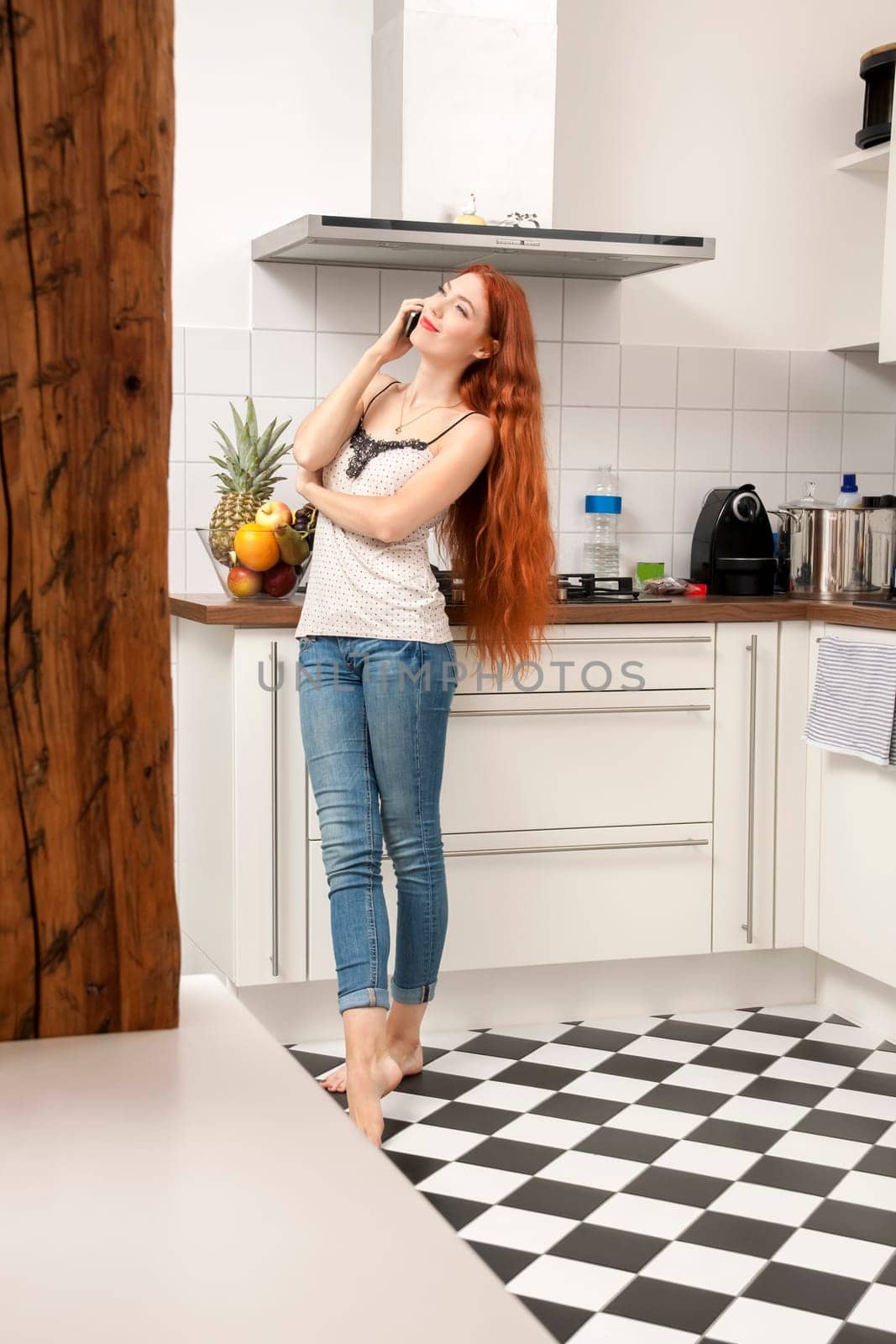 Happy young woman with mobile phone smartphone at home in the kitchen while making phone calls with long red hair