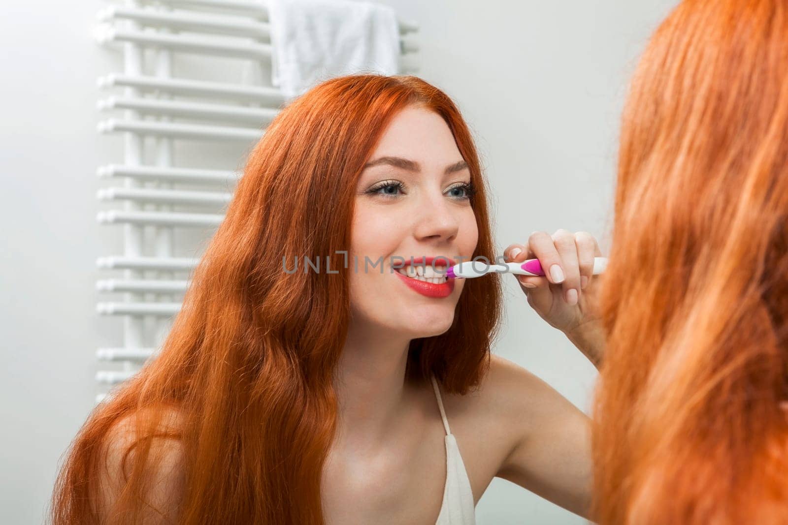 Attractive young woman in the morning brushing her teeth in the mirror in the bathroom with long red hair