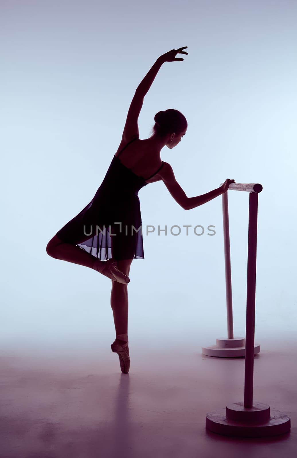 young ballerina stretching on the bar on blue background. The outline shooting - silhouette of girl
