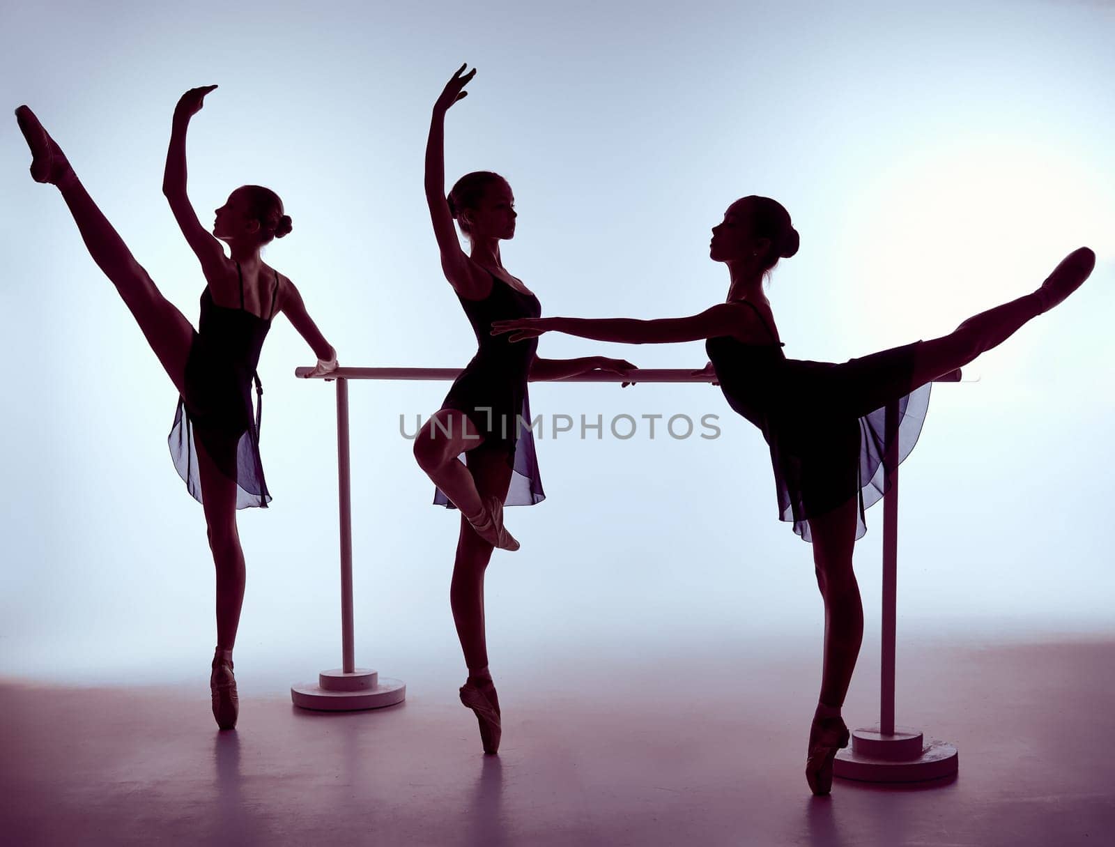 Three young ballerinas stretching on the bar on lilac background. The outline shooting - silhouettes of girls.