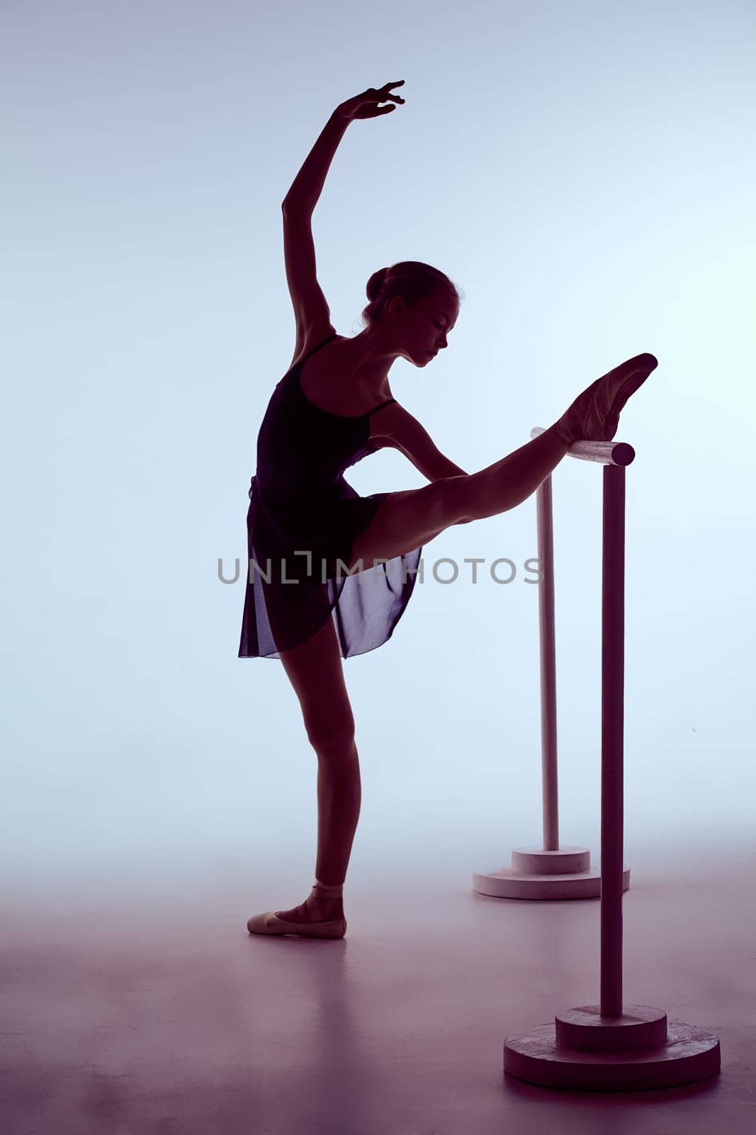 young ballerina stretching on the bar on blue background. The outline shooting - silhouette of girl