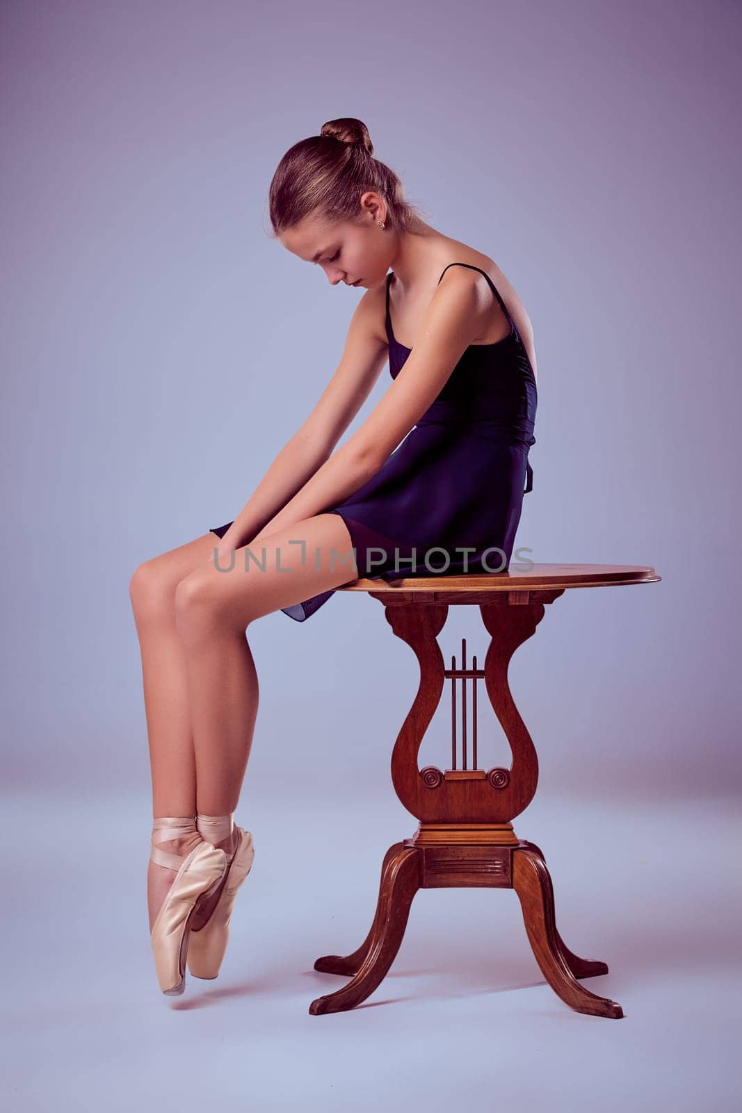 young ballerina in a blue dress sitting on wooden chair on a lilac background. Ballerina is wearing in pointe shoes