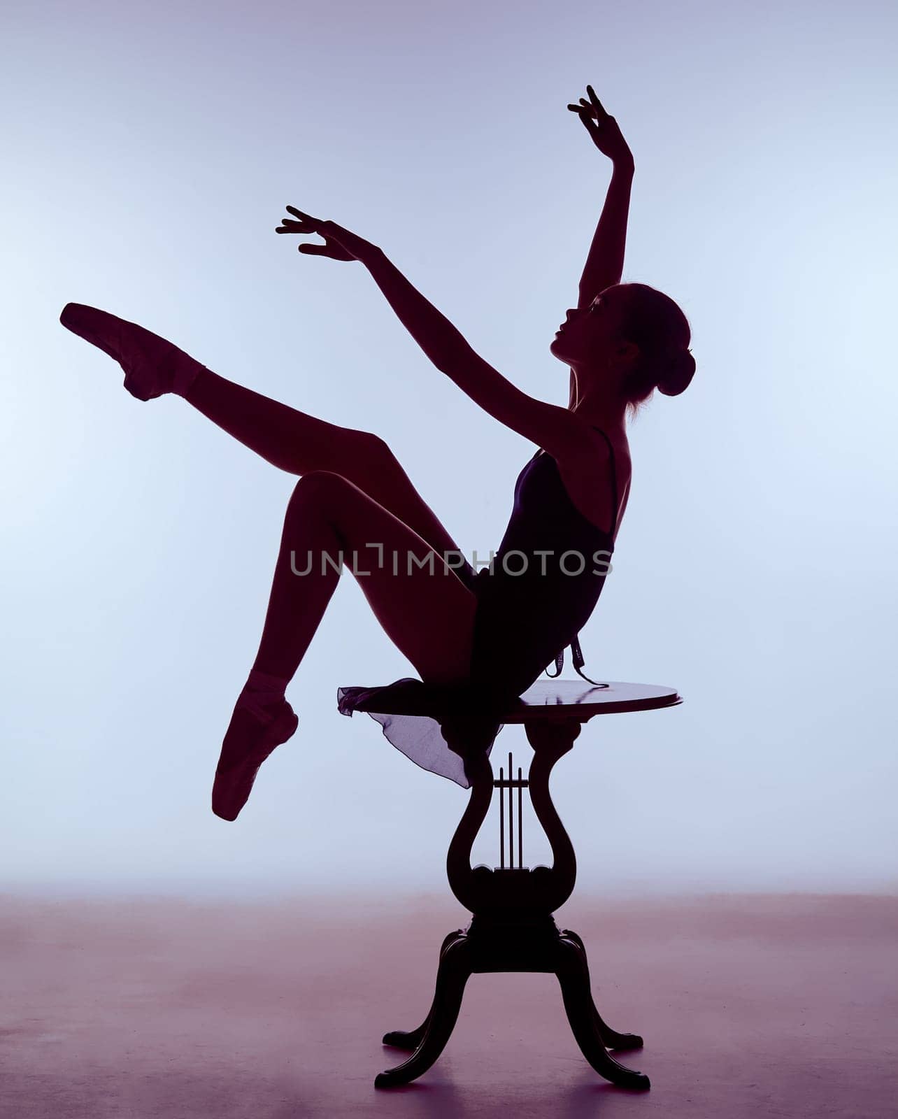 young ballerina in a blue dress sitting on wooden chair on a lilac background. Ballerina is wearing in pointe shoes.  The outline shooting - silhouette of girl