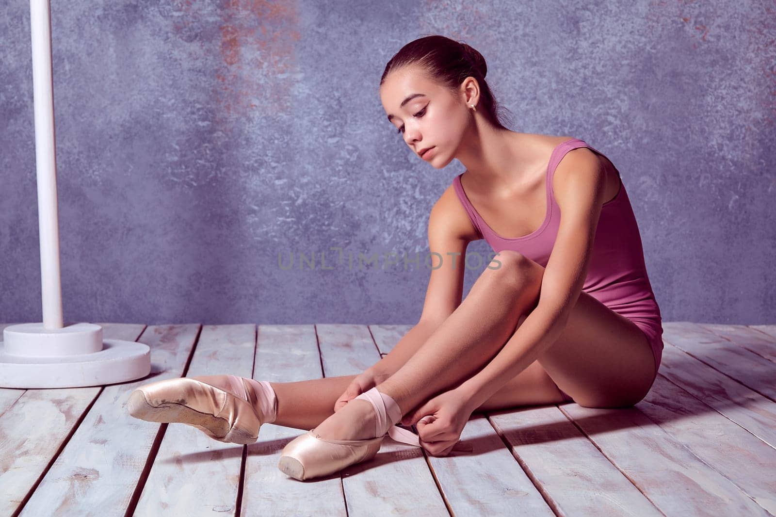 young ballerina putting on her ballet shoes on the wooden floor on a pink background