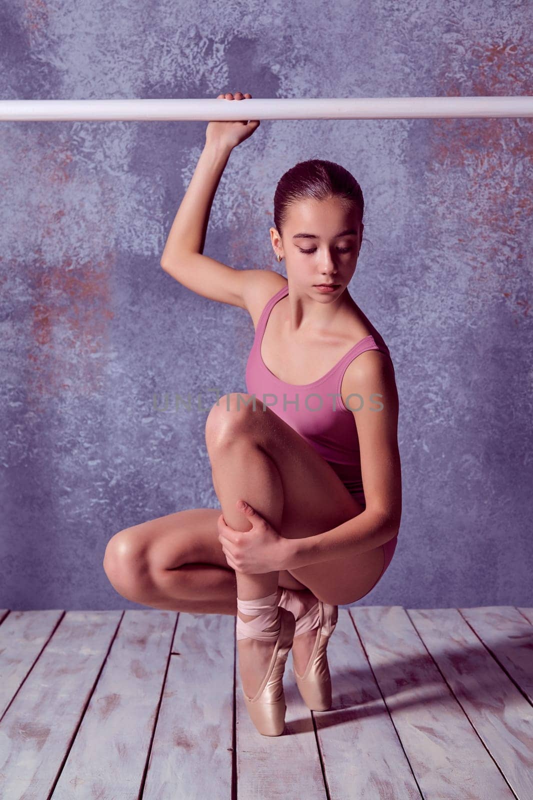 The young ballerina stretching on the bar on lilac background of wooden floor