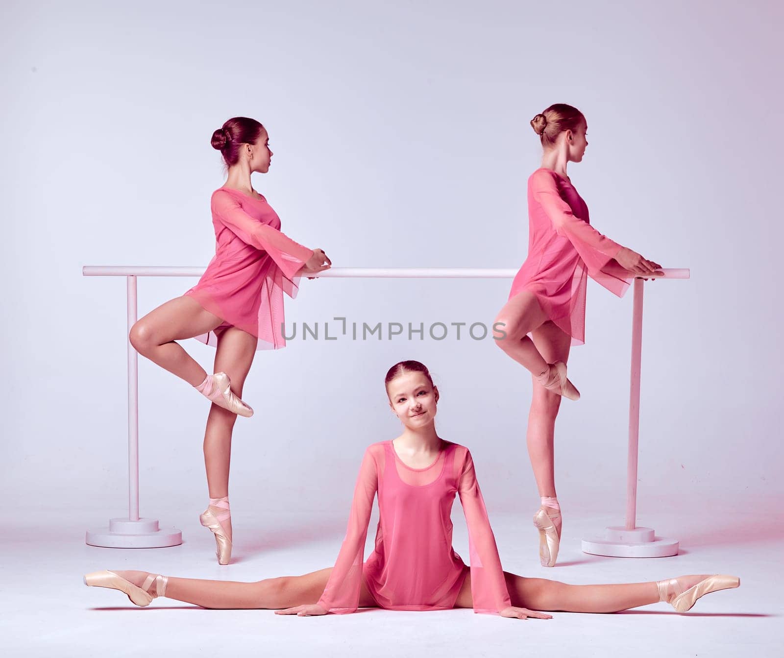 Three young ballerinas  in a pink dresses stretching on the bar on beige background