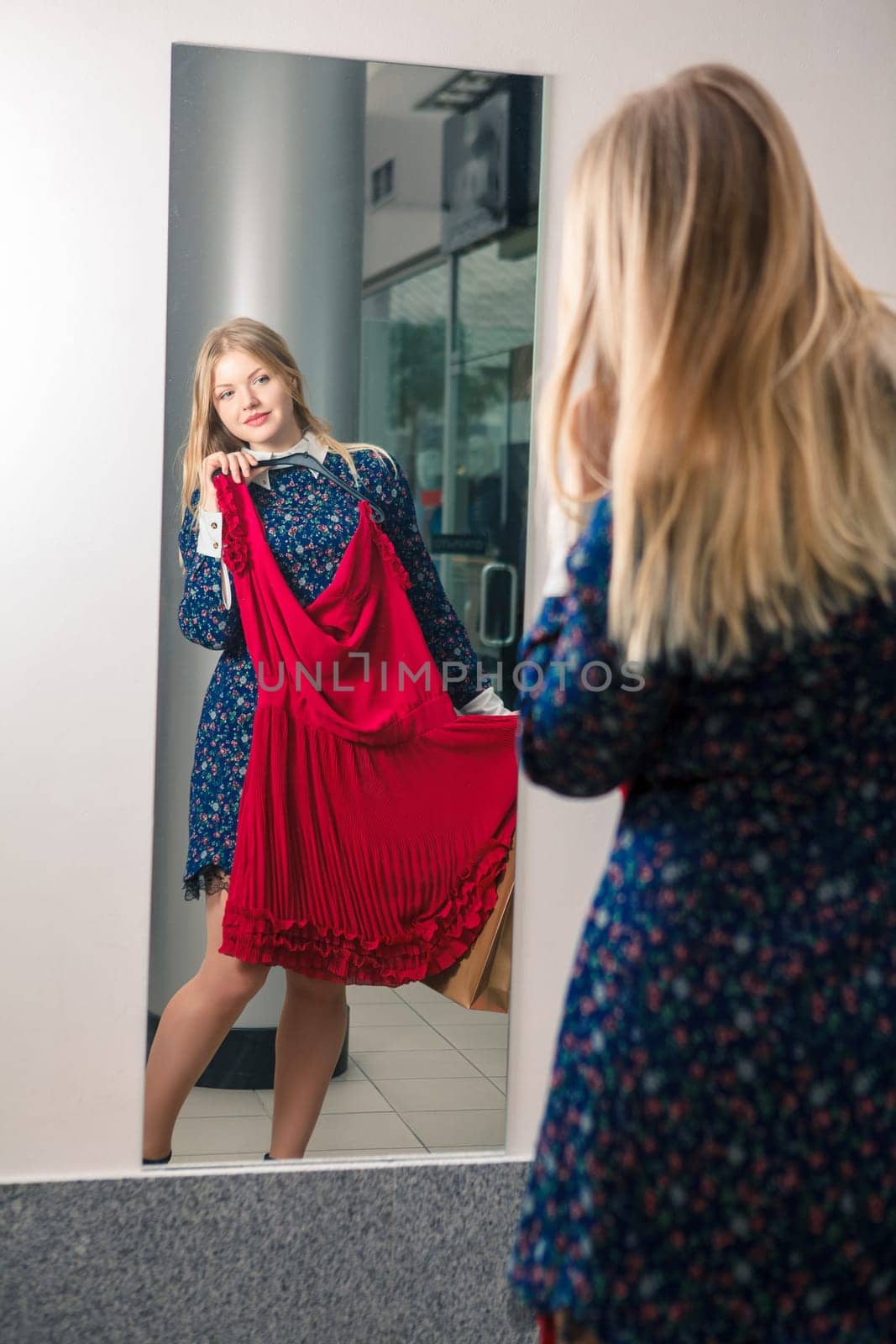 Woman trying red dress shopping for clothing. Beautiful happy blonde smiling young shopper woman looking in mirror standing in clothes store.