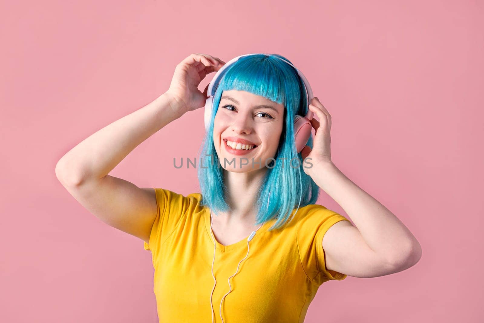 Beautiful smiling young woman with blue color dyed hair with pink headphones listening to music and dance in front of pastel studio background.