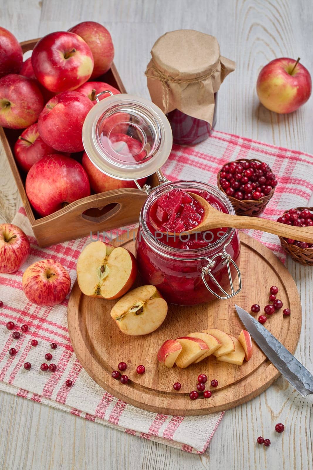 Still life with fresh lingonberries, red apples with knife on wooden cutting board and glass jar of homemade jam on light background. Autumn harvest, Home canning. Top view
