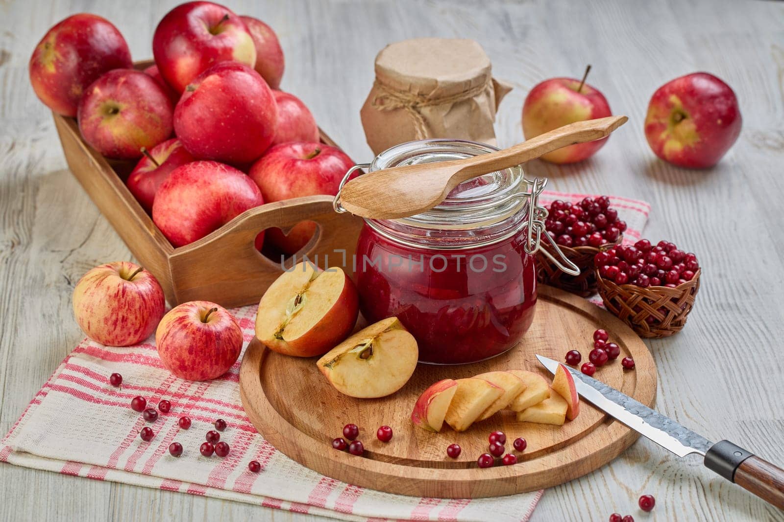 Still life with fresh lingonberries, red apples with knife on wooden cutting board and glass jar of homemade jam on light background. Autumn harvest, Home canning.