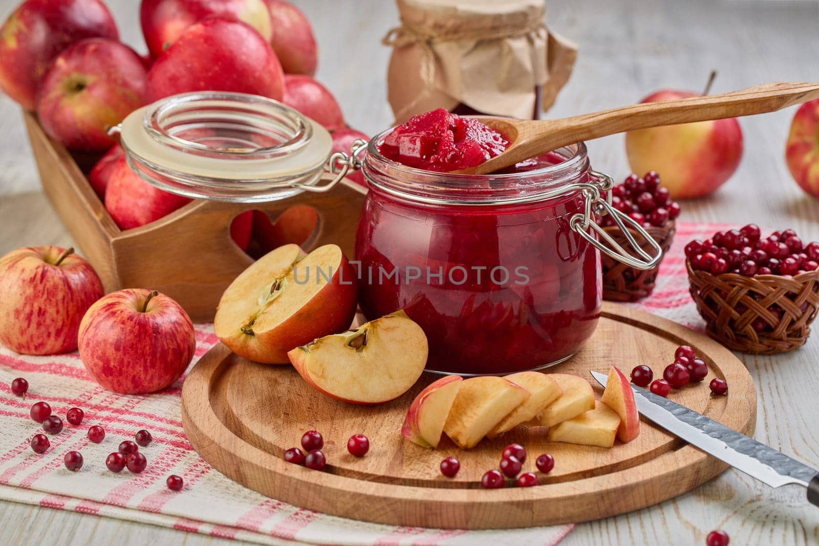 Still life with fresh lingonberries, red apples with knife on wooden cutting board and glass jar of homemade jam on light background. Autumn harvest, Home canning.