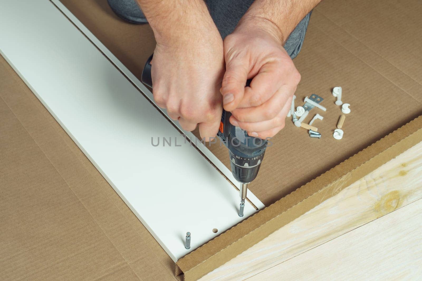 Male hands hold cordless electric screwdriver, screw to fix walls of white drawer, assembling dresser or wardrobe on floor. Self-assembly of furniture at home, DIY, furniture repair and rearrangement
