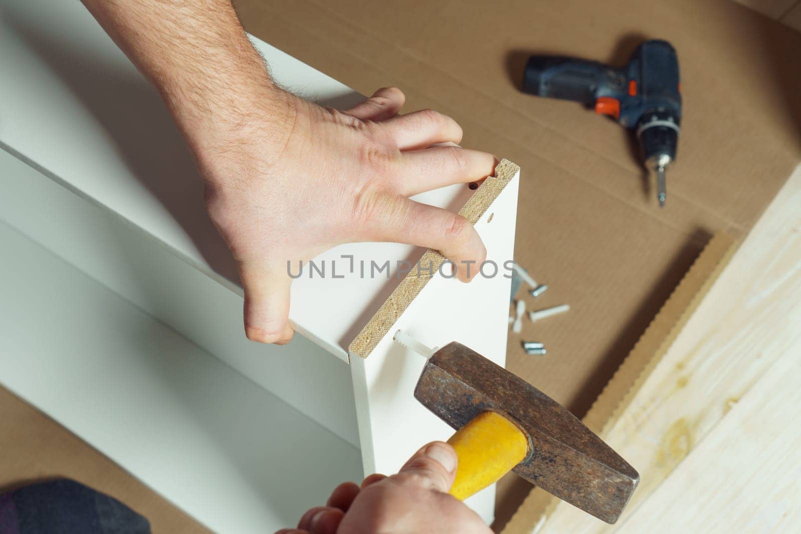 Male hands hold metal hammer with yellow plastic handle, driving dowel, assembling white drawer of dresser or wardrobe. Self-assembly of furniture at home, DIY, furniture repair and rearrangement