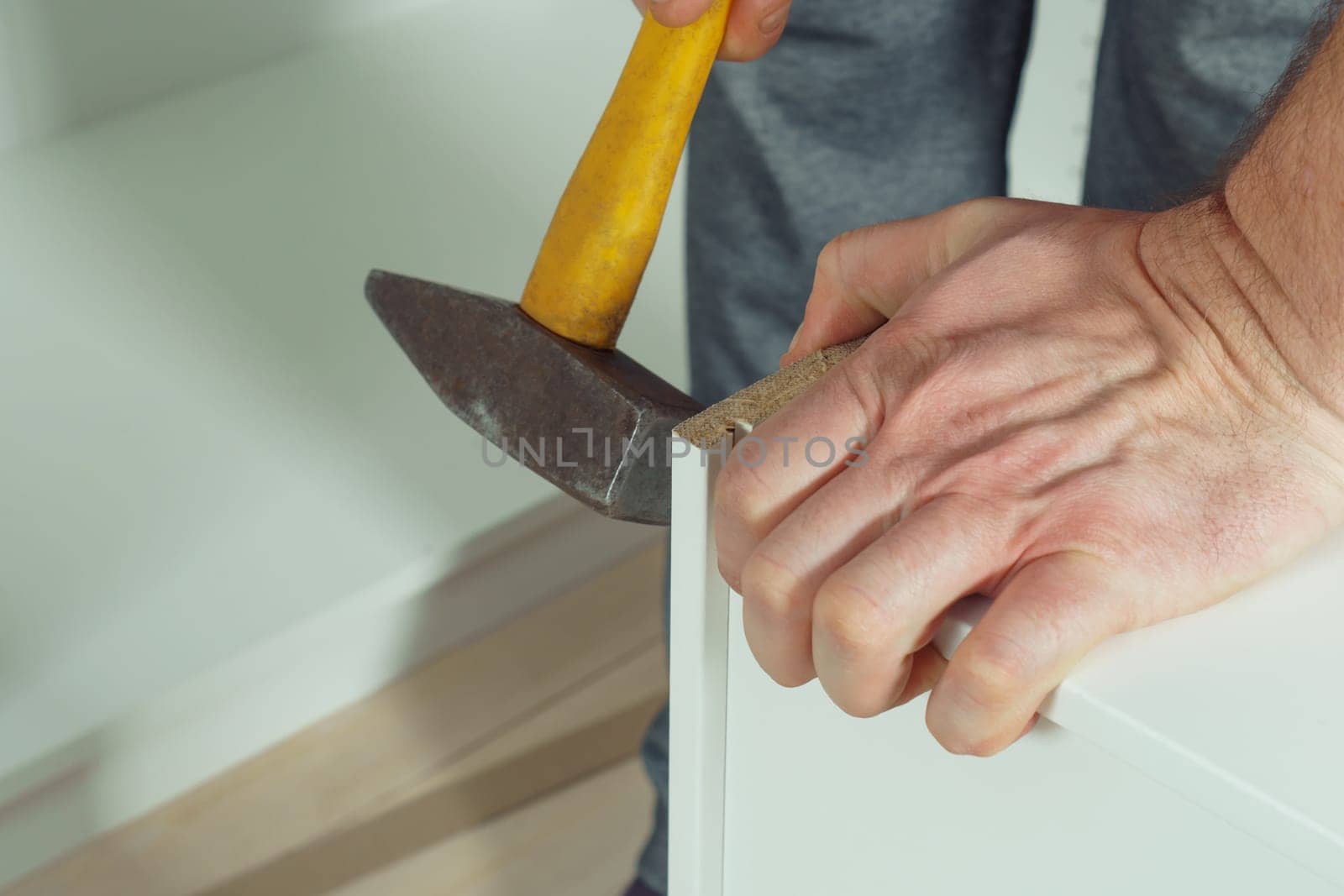 Male hands hold metal hammer with yellow plastic handle, driving dowel, assembling white drawer of dresser or wardrobe. Self-assembly of furniture at home, DIY, furniture repair and rearrangement