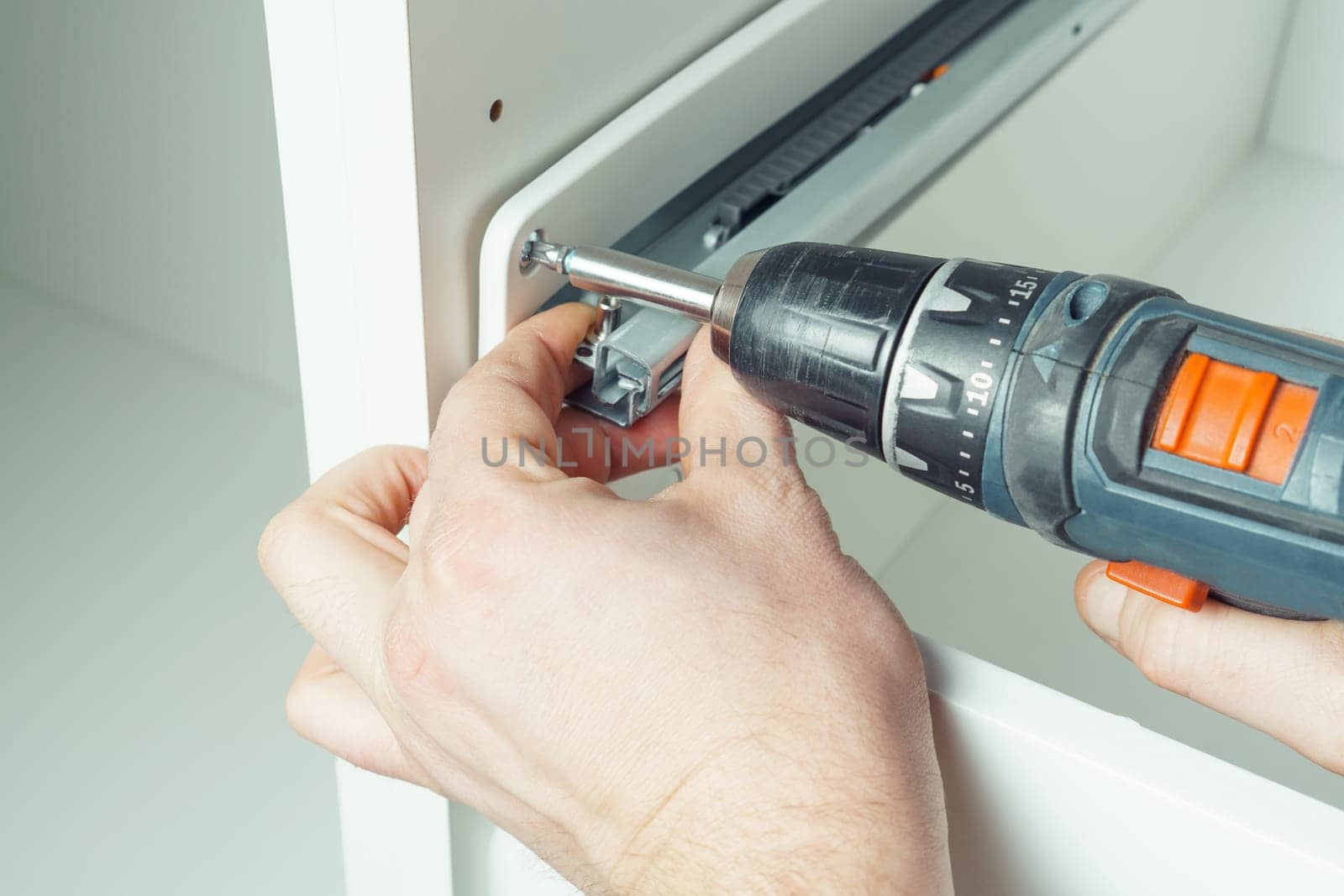 Male hands hold cordless electric screwdriver, screw to fix rail, assembling drawer of white dresser or wardrobe. Self-assembly of furniture at home, DIY, furniture repair and rearrangement