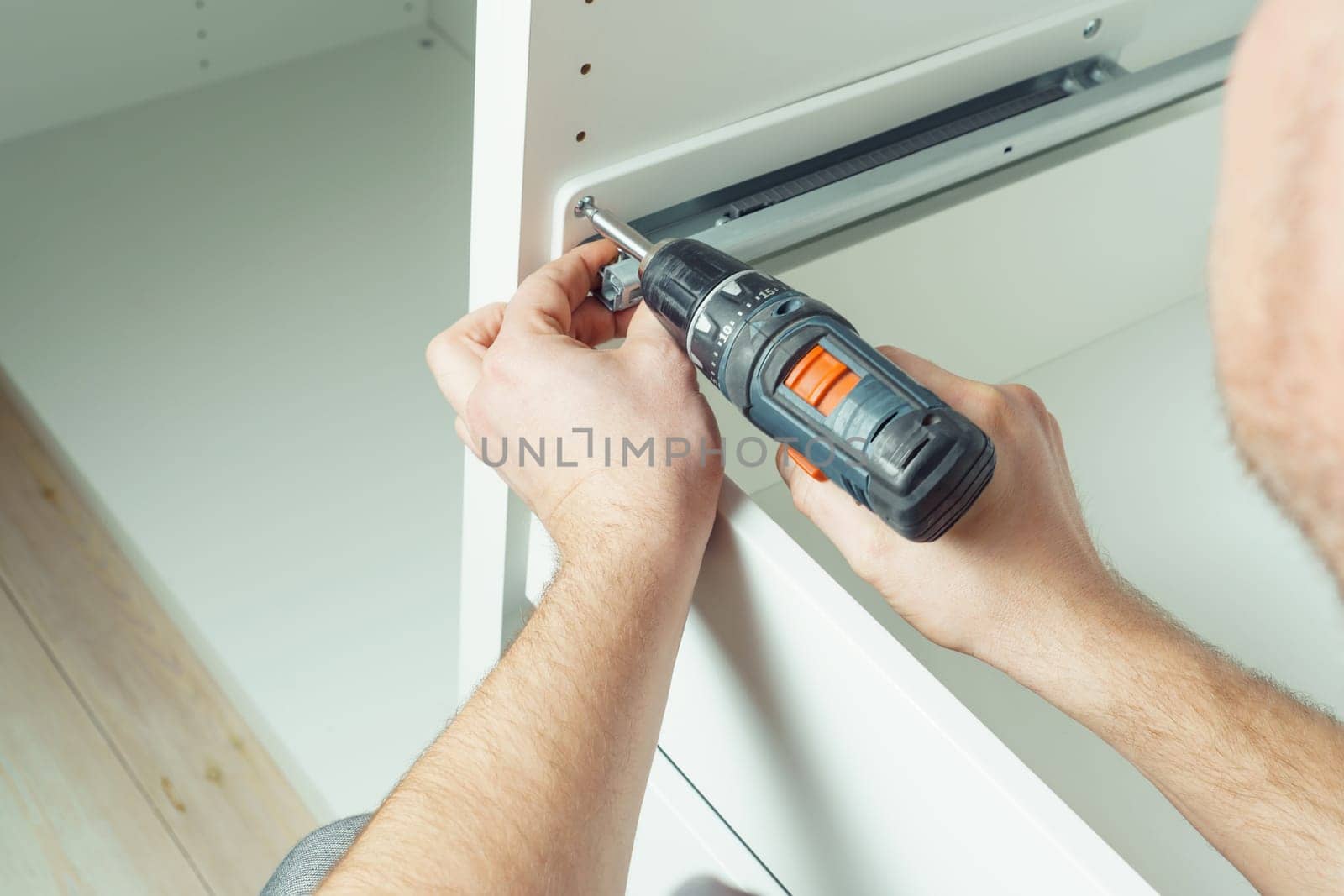 Male hands hold cordless electric screwdriver, screw to fix rail, assembling drawer of white dresser or wardrobe. Self-assembly of furniture at home, DIY, furniture repair and rearrangement