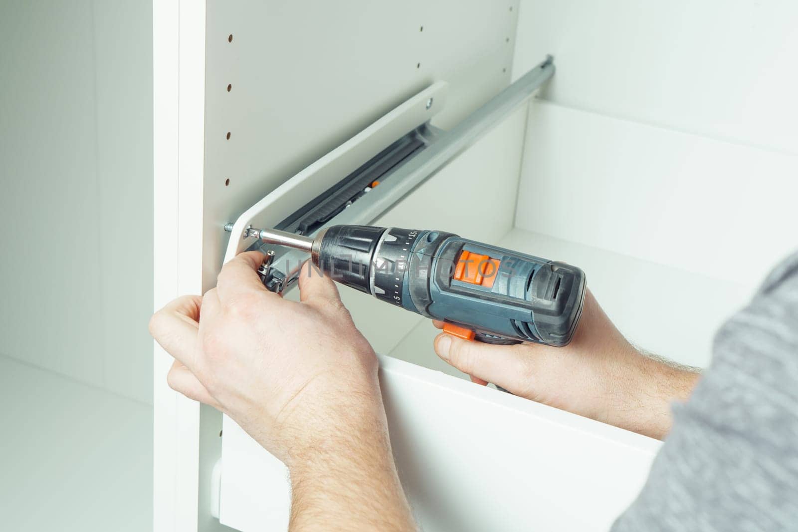 Male hands hold cordless electric screwdriver, screw to fix rail, assembling drawer of white dresser or wardrobe. Self-assembly of furniture at home, DIY, furniture repair and rearrangement