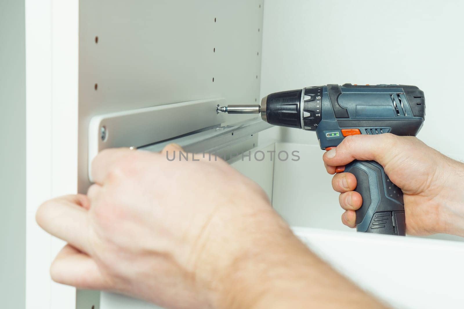 Male hands hold cordless electric screwdriver, screw to fix rail, assembling drawer of white dresser or wardrobe. Self-assembly of furniture at home, DIY, furniture repair and rearrangement