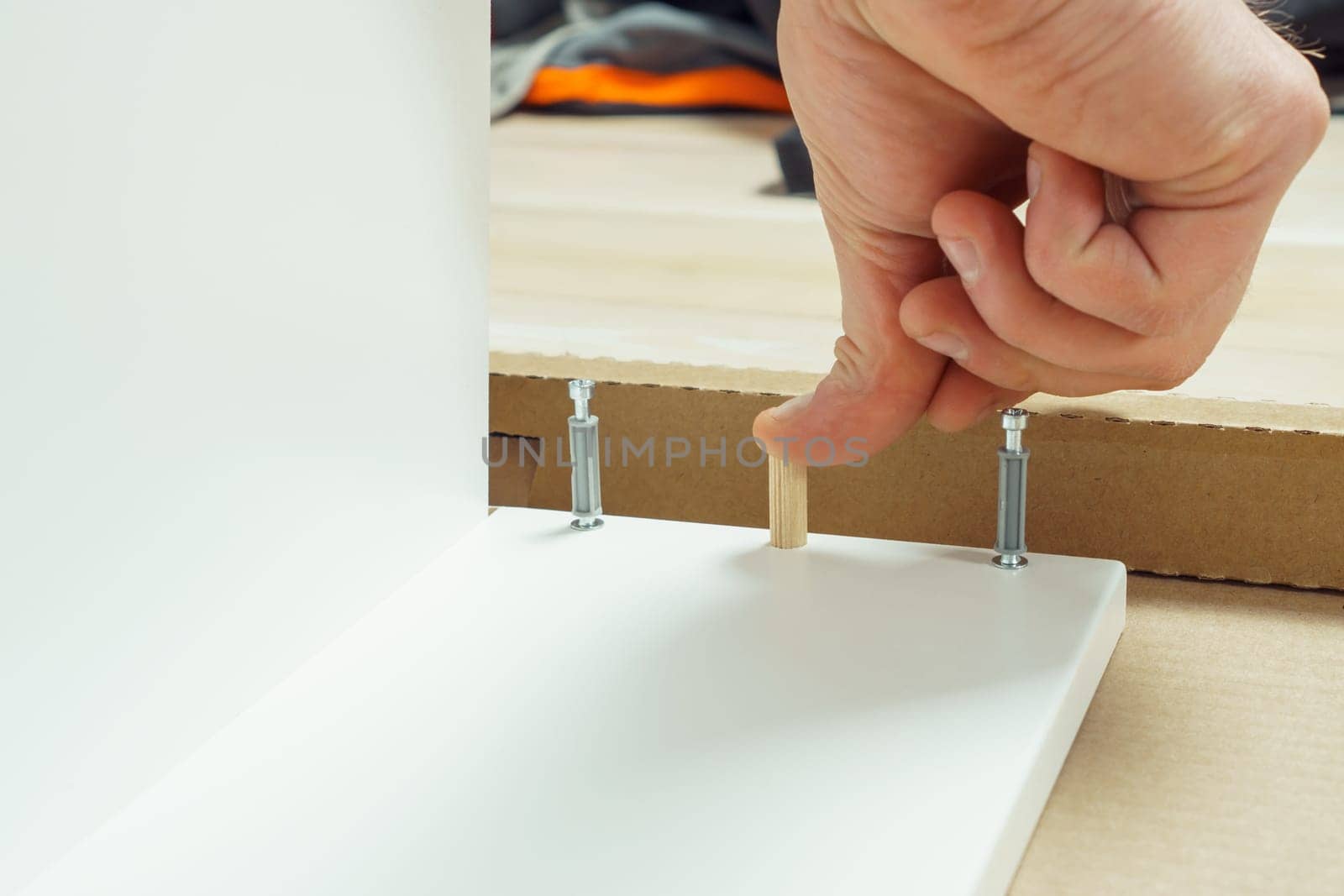 Male hands finger wooden dowel to fix walls of white drawer, assembling dresser or wardrobe on floor. Eccentric furniture ties on sides. Self-assembly of furniture at home, DIY, furniture repair