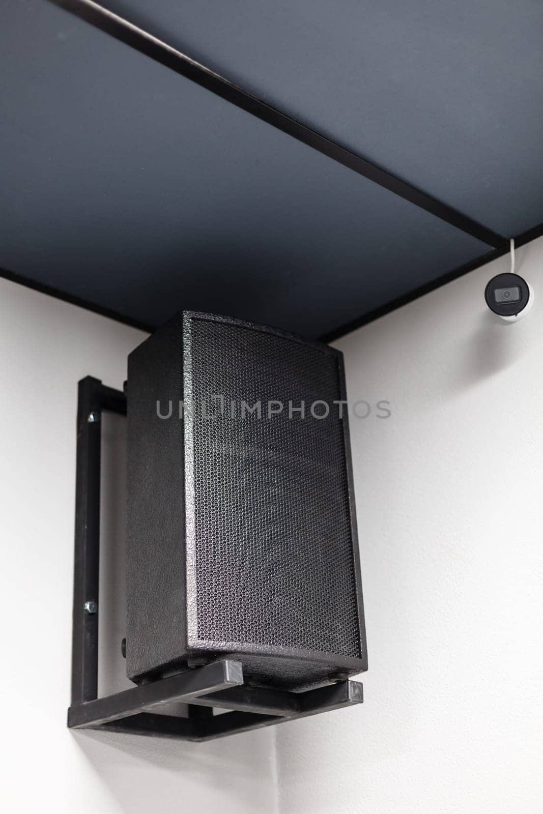 Close up of black big musical powerful speaker with protective grill hanging on wall in public place. Music speaker hangs on white wall near CCTV camera.