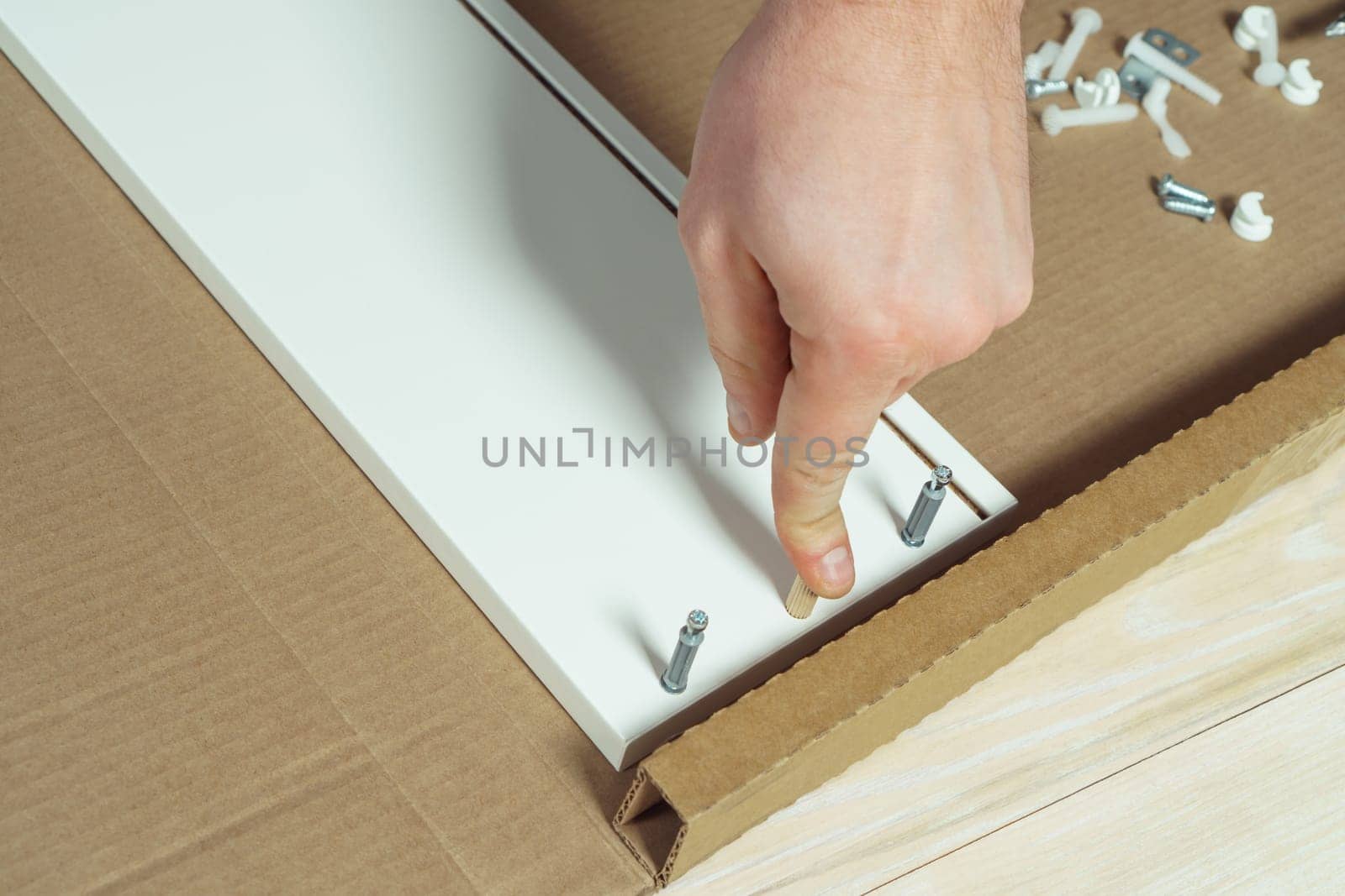 Male hands finger wooden dowel to fix walls of white drawer, assembling dresser or wardrobe on floor. Eccentric furniture ties on sides. Self-assembly of furniture at home, DIY, furniture repair