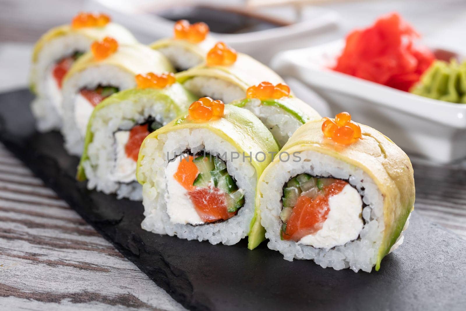 Close up of delicious pieces of sushi covered with fresh slices of avocado and red caviar. Sushi roll with salmon, cheese and cucumber inside stands on black metal board for serving sushi.
