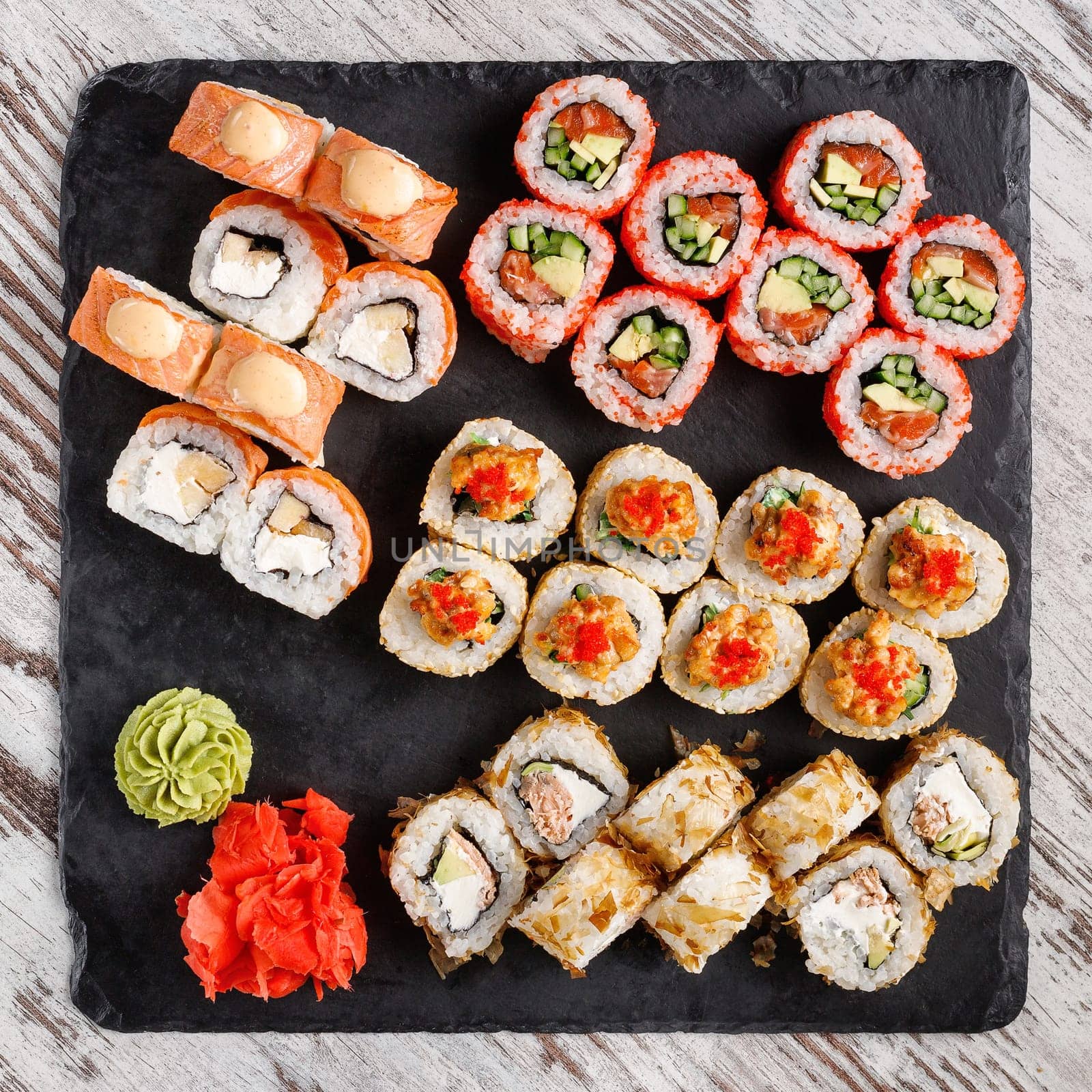Asian cuisine. Large sushi set of various sushi rolls is served on black square metal slate. Top view of different rolls with variety of fresh ingredients and wasabi with ginger on edge.
