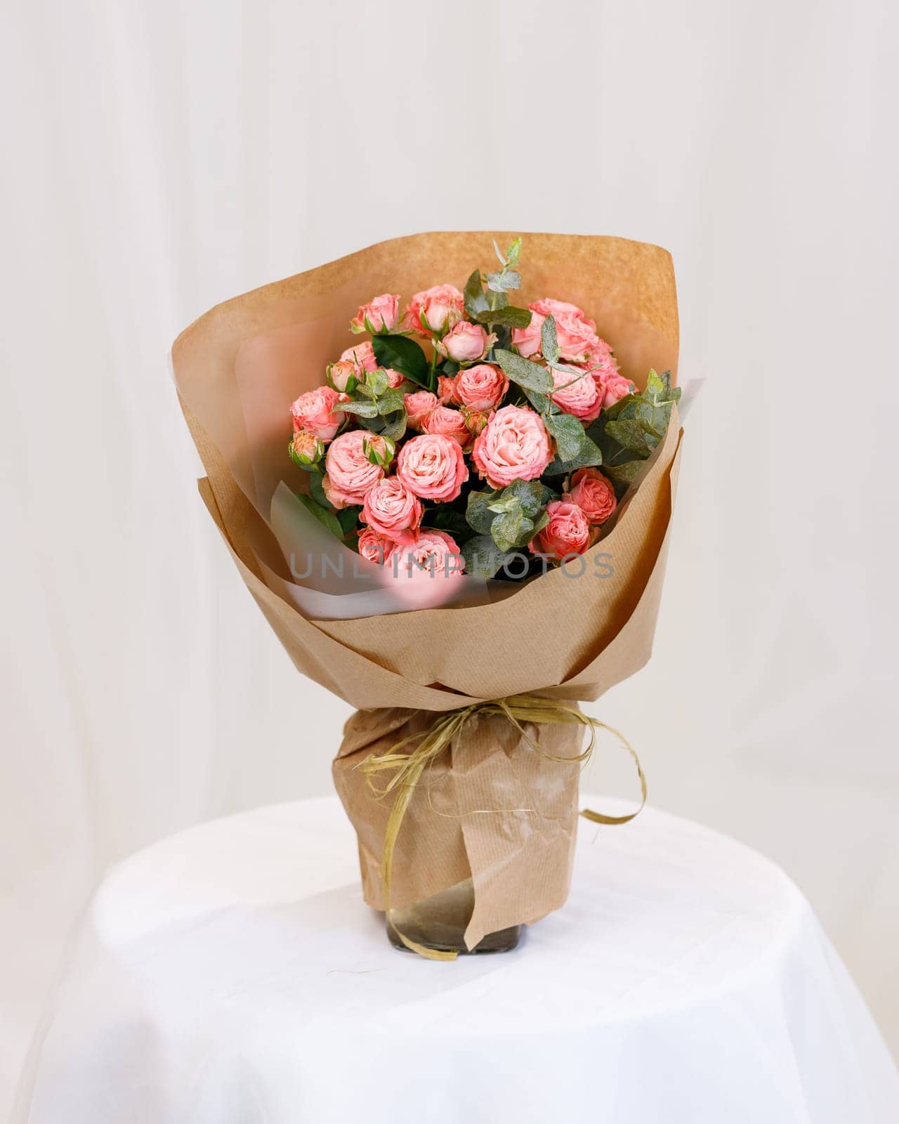 Bouquet of pink spray roses with sprigs of green eucalyptus in vase on stand on white textured background. Festive bouquet of flowers wrapped in brown kraft paper. Floral concept.